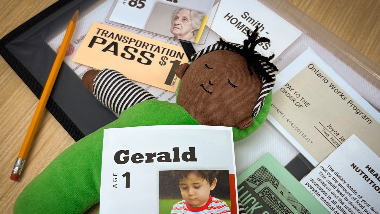 A participation kit from the poverty simulator is arranged on a table, featuring a doll, transport pass, Ontario Works program certificate and other materials in a binder.