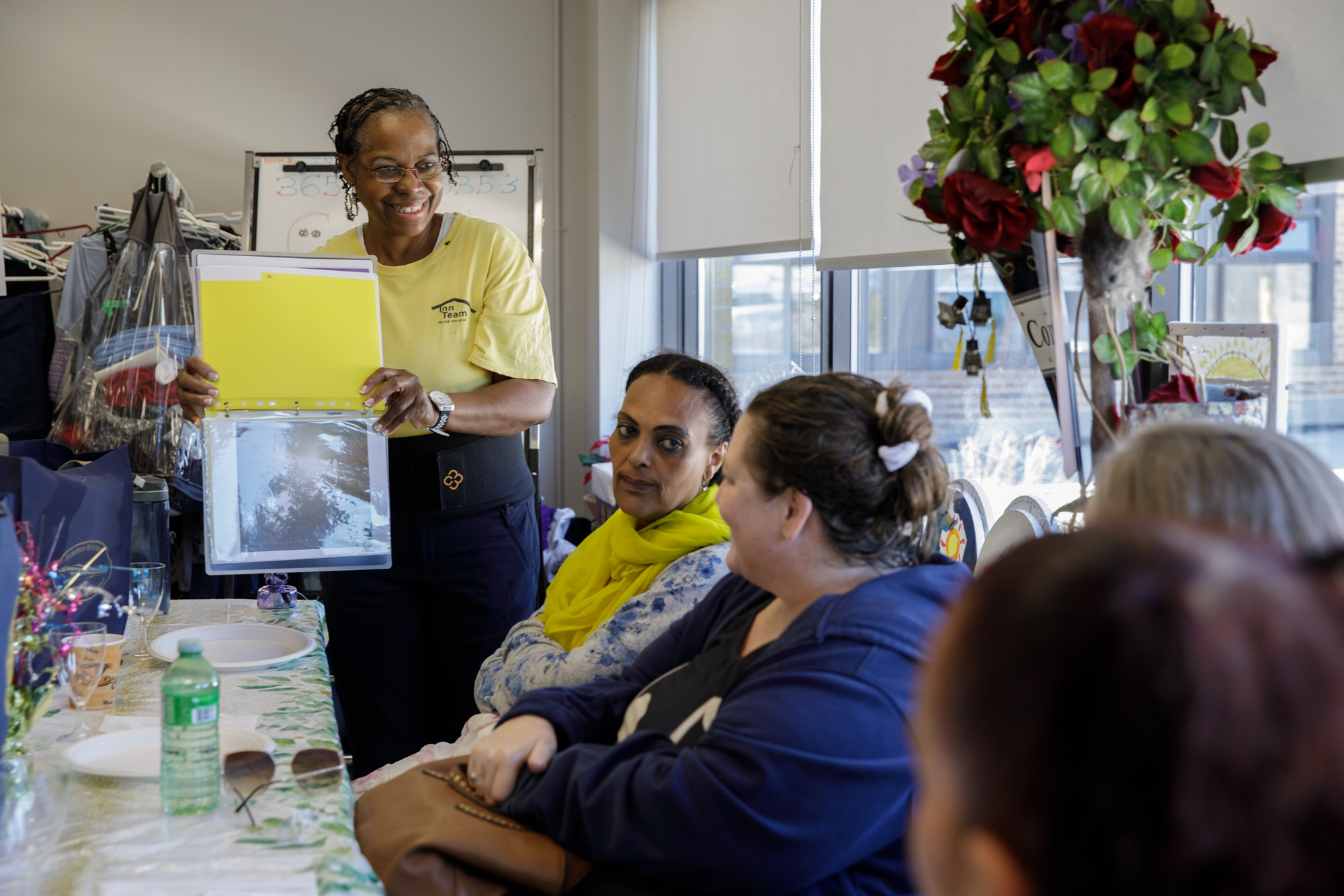 Carleen stands at table in a group setting, showing a binder of her photography to participants in the United Way-supported Getting Ahead program. People gathered around the table look on as she shares her work.