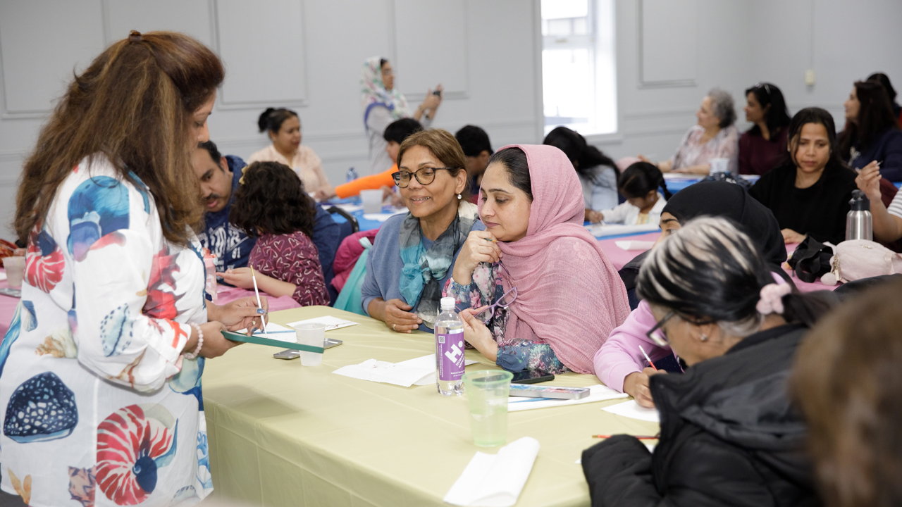 A group of women sit along a table with art supplies speaking to an instructor at a Seeds for Change Watercolour Workshop in Cooksville.