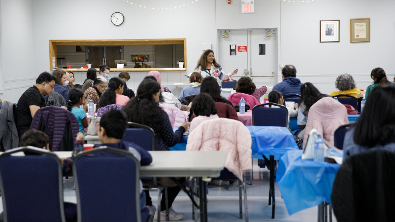A woman leads a group of people sitting at long tables at a Seeds for Change Watercolour Workshop in Cooksville.