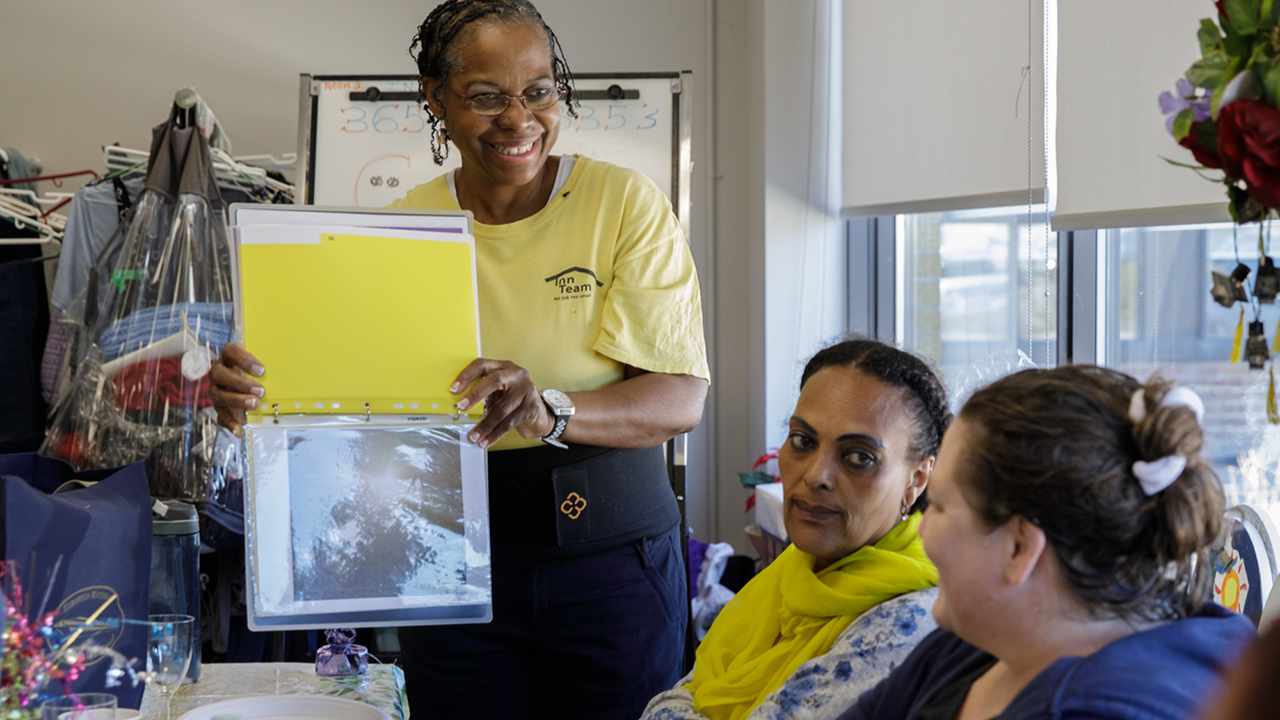 Carleen stands at table in a group setting, showing a binder of her photography to participants in the United Way-supported Getting Ahead program. People gathered around the table look on as she shares her work.