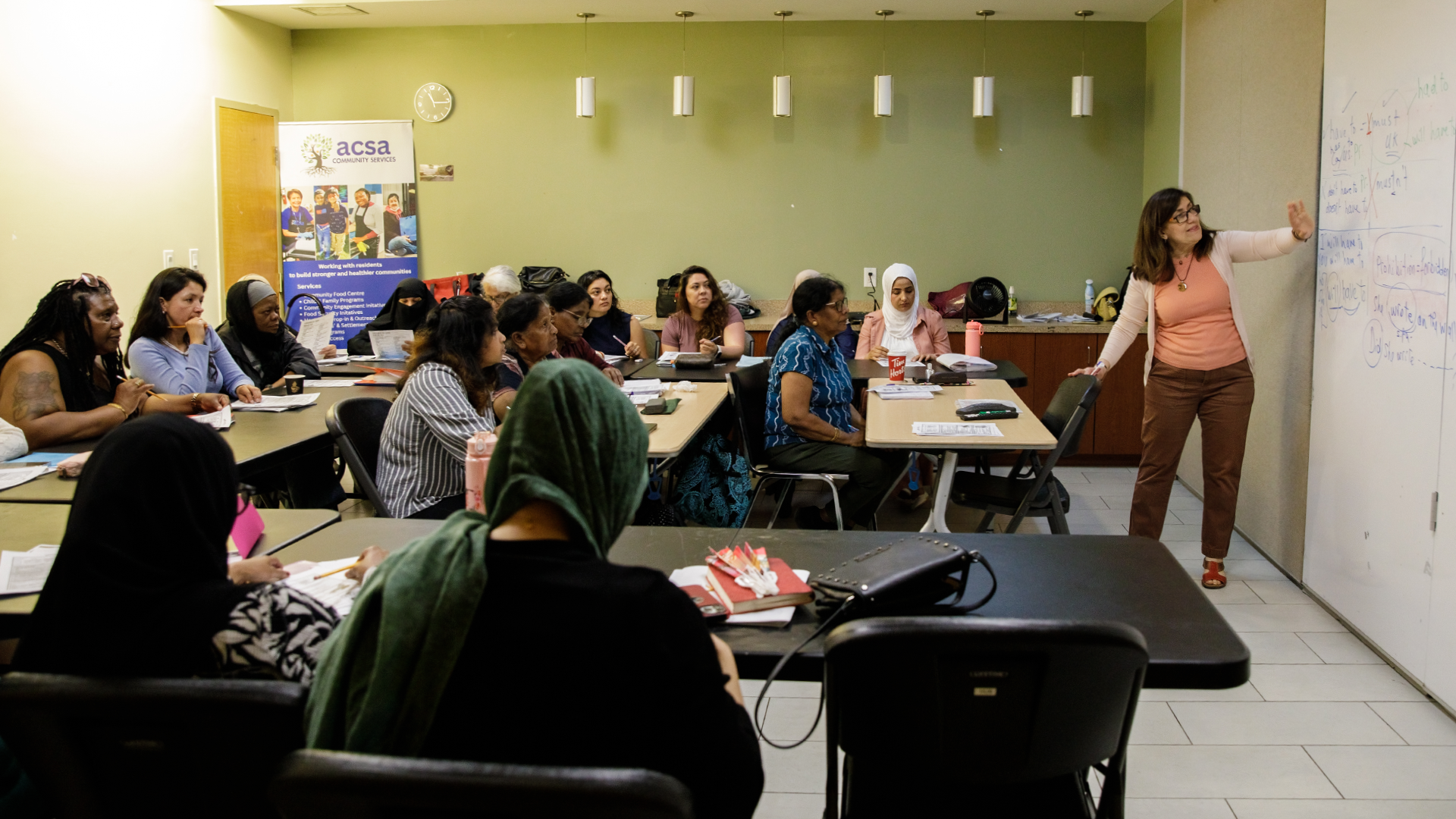 A woman teaches English to newcomers in a classroom at Dorset Park Community Hub, gesturing toward a whiteboard while speaking.