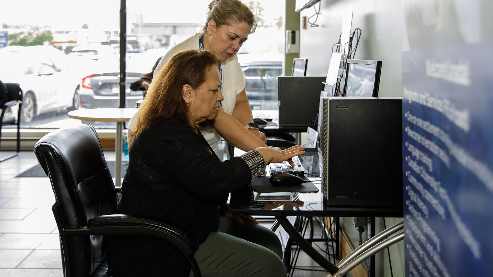 A woman is talking with another woman, while working on something on a computer.