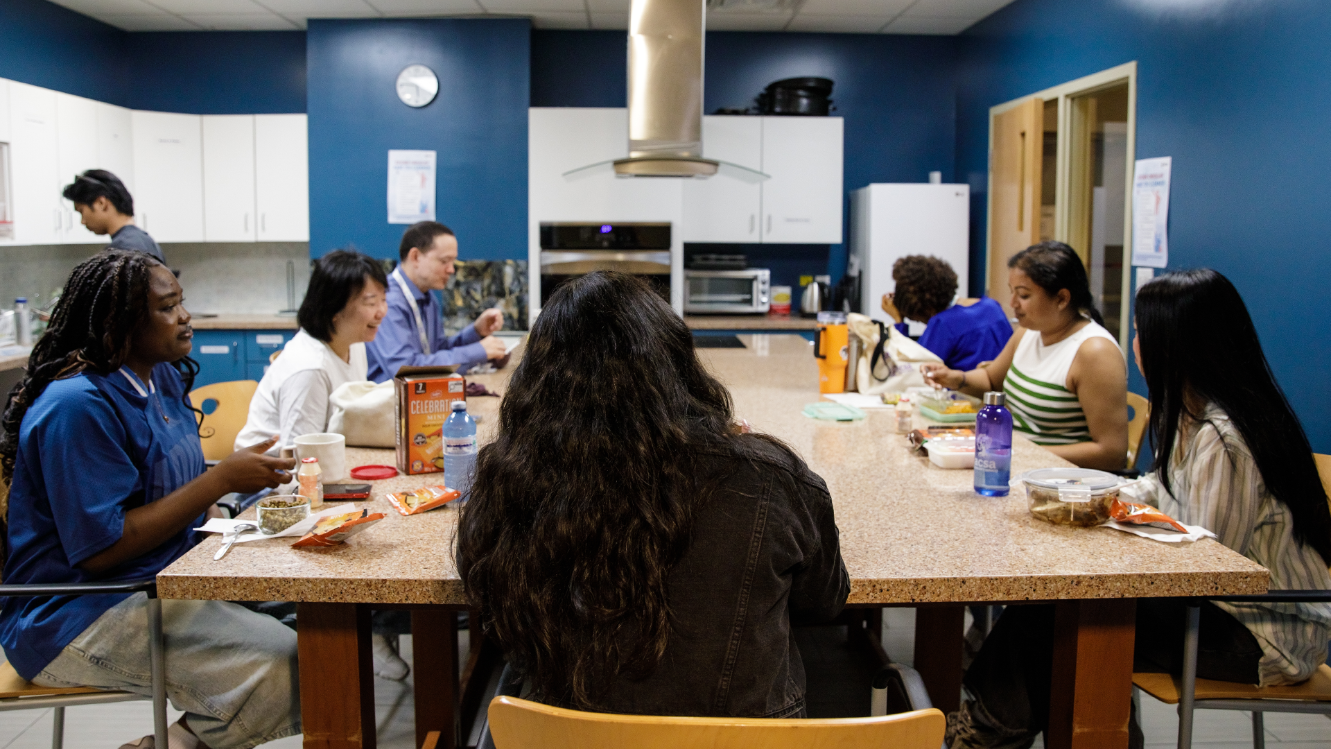A group of people eat together and talk, gathered around a table, during a lunch break.