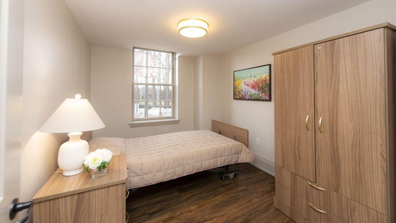 Warmly lit, clean bedroom with a side table, window, wardrobe, and wall art.