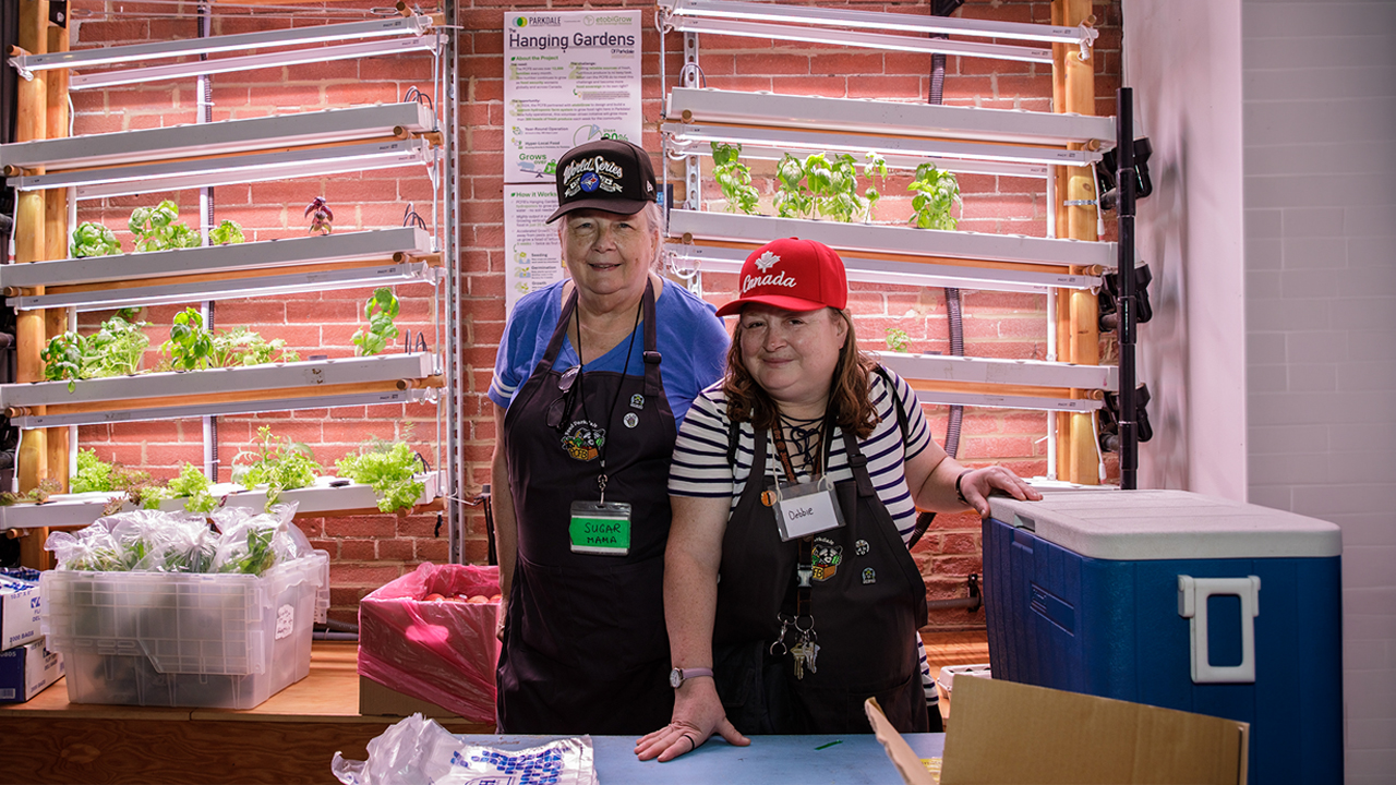 Two women wearing caps and aprons stand in front of a hydroponic wall at Parkdale Community Food Bank