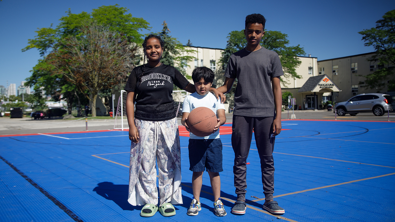 Three children of different ages stand on a multisport field. The child in the centre is holding a basketball.