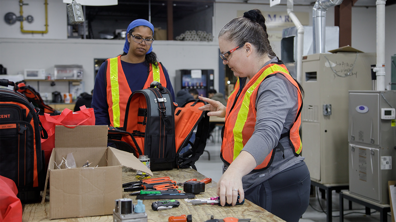Two participants in The Redwood's Women in Trades & Technology program wear reflective safety vests as they pack supplies into a bag at a worktable.